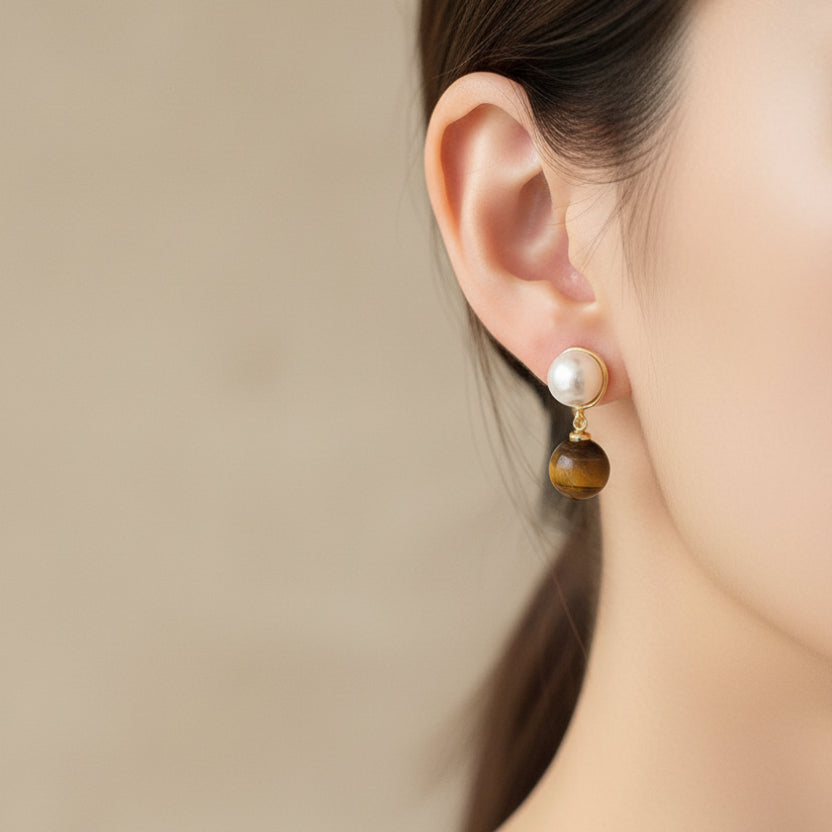 Close-up of an ear wearing a pearl and brown stone earring against a beige background